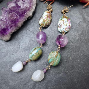 Può includere: Orecchini pendenti con conchiglie di abalone iridescenti, perline di ametista viola e pietre color turchese. Gli orecchini sono collegati con filo color oro e hanno perni color oro. Gli orecchini sono esposti su una superficie grigio scuro.