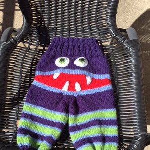 May include: Purple, green, and blue striped knitted pants with a monster face on the front. The monster has red teeth, white eyes, and a blue mouth.