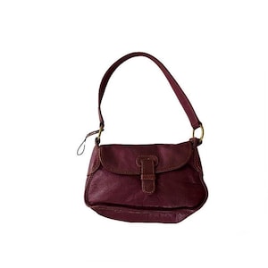 Könnte beinhalten: Eine burgunderfarbene Lederschultertasche mit Klappenverschluss und goldener Schnalle. Die Tasche hat einen kurzen Riemen und ist klein.