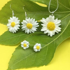May include: A set of daisy flower jewelry, including a necklace and earrings. The necklace has a single daisy pendant, while the earrings are a pair of dangle earrings with daisy charms. The jewelry is displayed on a green leaf against a yellow background.