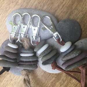 Puede incluir: Cuatro llaveros plateados con piedras grises y blancas apiladas unidas a correas de cuero marrón. Las piedras están apiladas en forma de pirámide y los llaveros están sobre una superficie de madera.