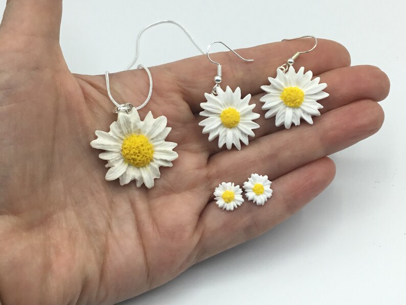 May include: A set of daisy flower jewelry, including a necklace, dangle earrings, and stud earrings. The flowers are white with yellow centers.