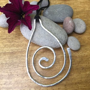 May include: A silver pendant necklace with a hammered metal spiral design. The pendant is hanging from a black cord and is displayed on a wooden surface with stones.