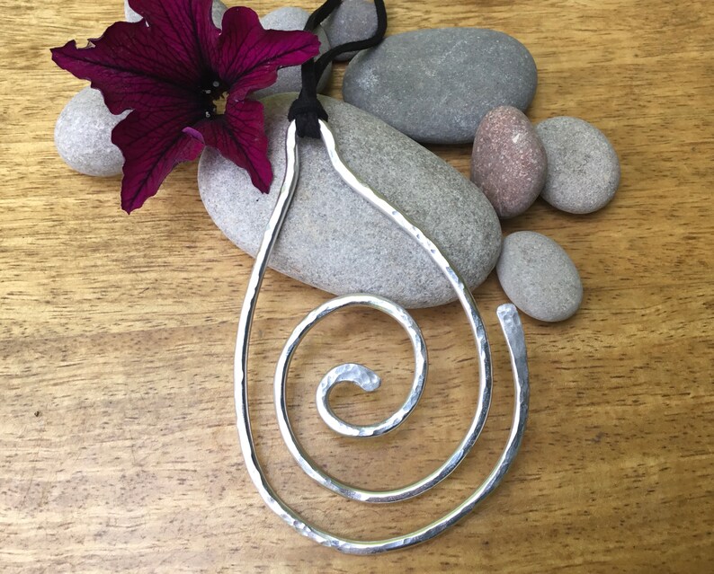 May include: A silver pendant necklace with a hammered metal spiral design. The pendant is hanging from a black cord and is displayed on a wooden surface with stones.