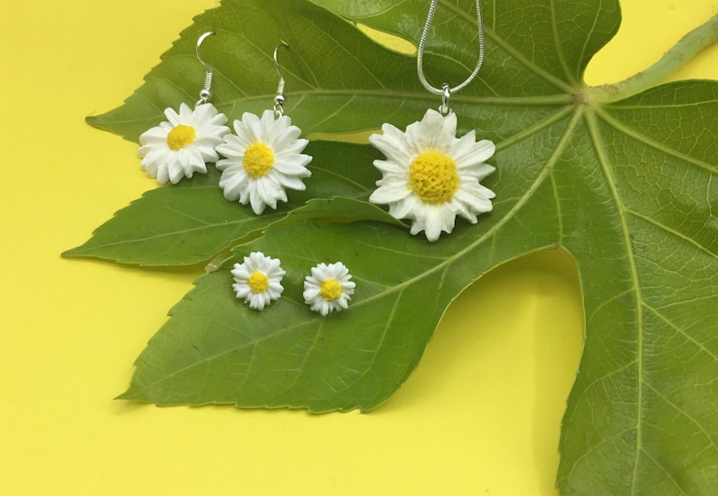 May include: A set of white daisy earrings and a necklace with a larger daisy pendant. The jewelry is displayed on a green leaf against a yellow background.