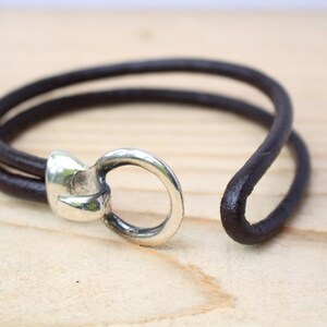 May include: A dark brown leather bracelet with a silver-colored clasp. The bracelet features a simple, minimalist design with two leather strands and a circular clasp. The bracelet is shown on a light wooden surface.