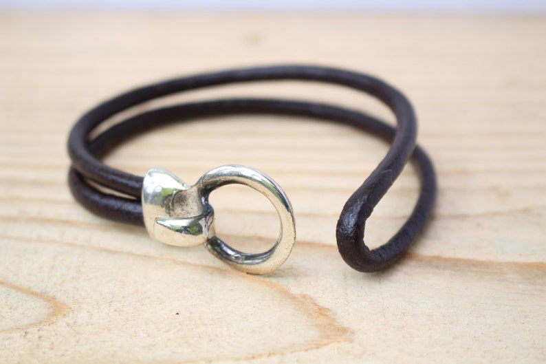 May include: A dark brown leather bracelet with a silver-colored clasp. The bracelet features a simple, minimalist design with two leather strands and a circular clasp. The bracelet is shown on a light wooden surface.