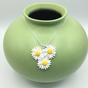 May include: A silver chain necklace with three white daisy flower charms with yellow centers. The necklace is displayed on a green ceramic vase.