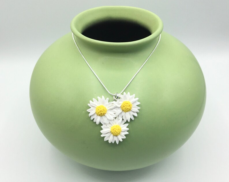 May include: A silver chain necklace with three white daisy flower charms with yellow centers. The necklace is displayed on a green ceramic vase.