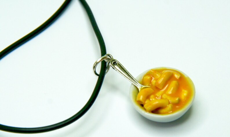May include: A black cord necklace with a silver spoon and a small white bowl filled with yellow macaroni cheese.