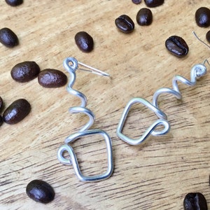 May include: A pair of silver wire earrings shaped like coffee cups. The earrings are on a wooden surface with coffee beans scattered around them.