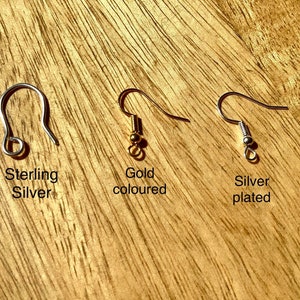 May include: Three earring hooks on a wooden surface. The hook on the left is sterling silver. The middle hook is gold-colored. The hook on the right is silver-plated.