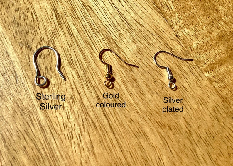 May include: Three earring hooks on a wooden surface. The hook on the left is sterling silver. The middle hook is gold-colored. The hook on the right is silver-plated.