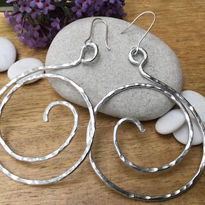 May include: Silver hoop earrings with a hammered finish and a spiral design. The earrings are large and have a simple, modern style.