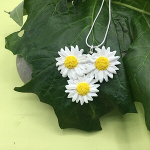 May include: A silver chain necklace with three white daisy flower charms. The flowers have yellow centers and are arranged in a cluster.