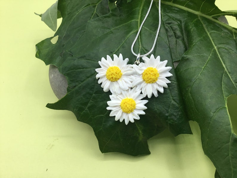 May include: A silver chain necklace with three white daisy flower charms. The flowers have yellow centers and are arranged in a cluster.