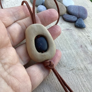 Puede incluir: Un collar de cordón de cuero marrón con un colgante de piedra beige. La piedra tiene una piedra ovalada de color rojo oscuro engastada en su interior.