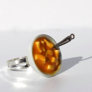 Puede incluir: Un anillo de cer&aacute;mica blanca con un taz&oacute;n en miniatura de sopa naranja y una cuchara de plata. El taz&oacute;n est&aacute; lleno de peque&ntilde;os trozos de comida blancos y naranjas.