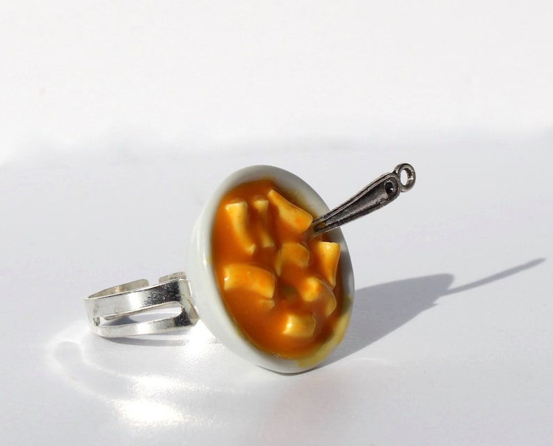 Puede incluir: Un anillo de cer&aacute;mica blanca con un taz&oacute;n en miniatura de sopa naranja y una cuchara de plata. El taz&oacute;n est&aacute; lleno de peque&ntilde;os trozos de comida blancos y naranjas.