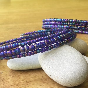 May include: A multi-strand beaded bracelet with iridescent blue beads wrapped around a silver wire. The bracelet is on a light brown wooden surface with two gray stones.