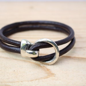 May include: A dark brown leather bracelet with a silver-colored clasp. The bracelet features two leather bands that loop around a circular clasp. The clasp is a prominent feature, adding a touch of shine to the accessory.