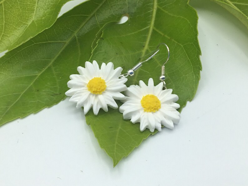 May include: A pair of white daisy earrings with yellow centres. The earrings are hanging from silver hooks.