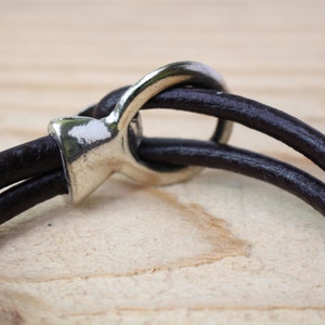 May include: Close-up of a dark brown leather bracelet with a silver-colored clasp. The bracelet features two leather bands that loop through the clasp. The background is a light brown wooden surface.