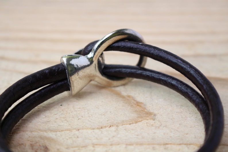 May include: Close-up of a dark brown leather bracelet with a silver-colored clasp. The bracelet features two leather bands that loop through the clasp. The background is a light brown wooden surface.