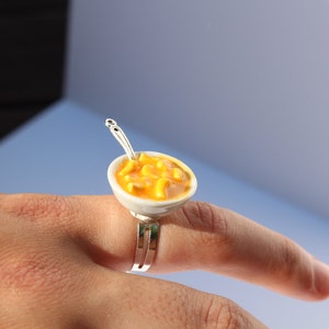 Puede incluir: Un anillo de plata con un cuenco de cer&aacute;mica blanca lleno de macarrones con queso amarillo. El cuenco tiene una peque&ntilde;a cuchara de plata unida al lado.