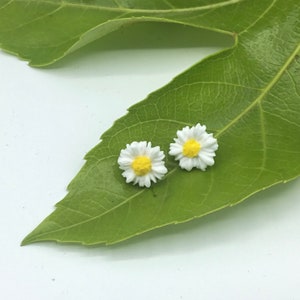 May include: A pair of white daisy earrings with yellow centres. The earrings are on a green leaf.