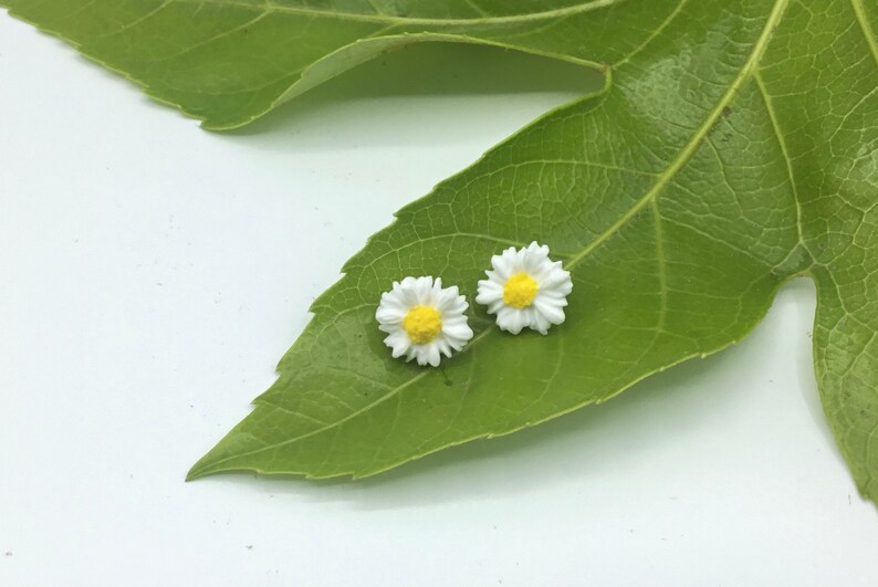 May include: A pair of white daisy earrings with yellow centres. The earrings are on a green leaf.