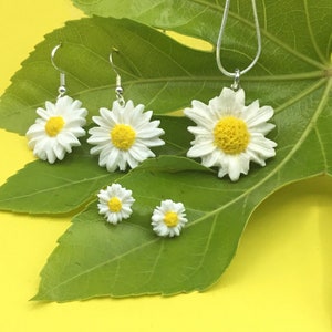 May include: A set of daisy flower jewelry, including a necklace, earrings, and a pair of stud earrings. The jewelry is made of white resin with yellow centers and silver accents. The jewelry is displayed on a green leaf against a yellow background.