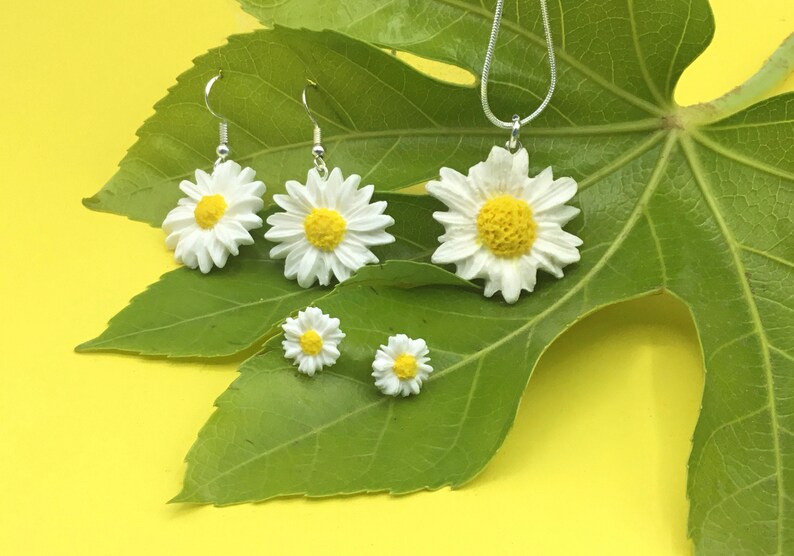 May include: A set of daisy flower jewelry, including a necklace, earrings, and a pair of stud earrings. The jewelry is made of white resin with yellow centers and silver accents. The jewelry is displayed on a green leaf against a yellow background.