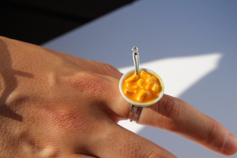 Puede incluir: Un anillo de plata con un cuenco de cer&aacute;mica blanca lleno de macarrones con queso naranja. El cuenco tiene una peque&ntilde;a cuchara de plata unida al lado.
