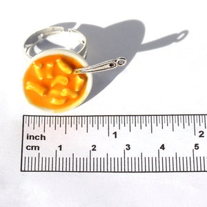 Puede incluir: Un anillo de plata con un cuenco blanco lleno de pasta naranja y una cuchara de plata. El anillo tiene un di&aacute;metro de aproximadamente 2,5 cm.