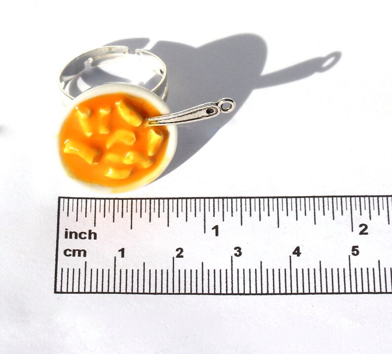 Puede incluir: Un anillo de plata con un cuenco blanco lleno de pasta naranja y una cuchara de plata. El anillo tiene un di&aacute;metro de aproximadamente 2,5 cm.