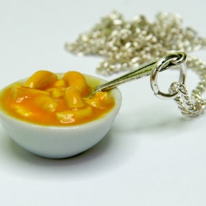 May include: A silver necklace featuring a miniature white bowl filled with orange and yellow macaroni cheese. The bowl contains a small silver spoon.