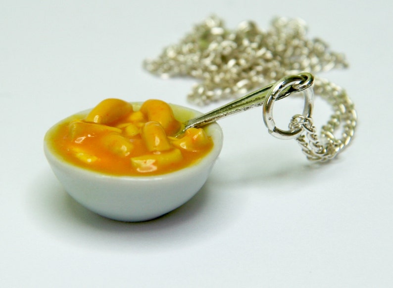 May include: A silver necklace featuring a miniature white bowl filled with orange and yellow macaroni cheese. The bowl contains a small silver spoon.