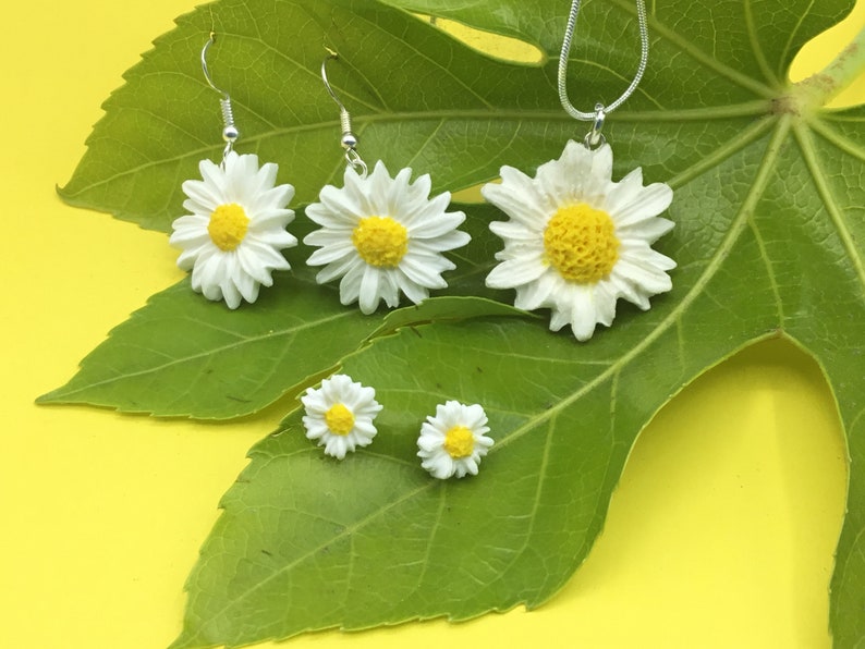 May include: A set of daisy flower jewellery, including a necklace and earrings. The necklace has a single daisy pendant, while the earrings are a pair of dangle earrings with daisy charms. The jewellery is displayed on a green leaf against a yellow background.