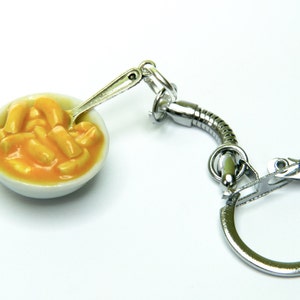 May include: A silver keychain featuring a miniature white bowl filled with yellow macaroni cheese. A tiny silver spoon is inside the bowl.