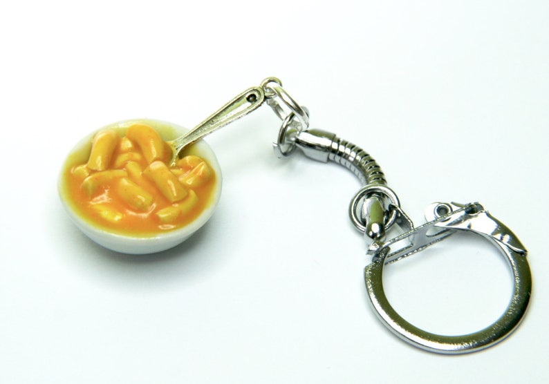 May include: A silver keychain featuring a miniature white bowl filled with yellow macaroni cheese. A tiny silver spoon is inside the bowl.