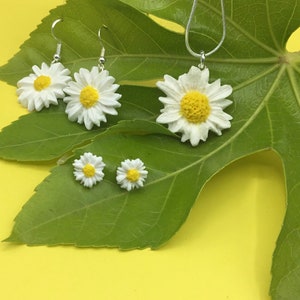 May include: A set of white daisy earrings and a necklace with a larger daisy pendant. The jewellery is displayed on a green leaf against a yellow background.