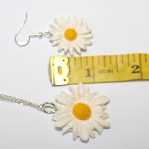 May include: A white daisy necklace and earrings set with a yellow centre. The necklace is on a silver chain and the earrings have silver hooks.