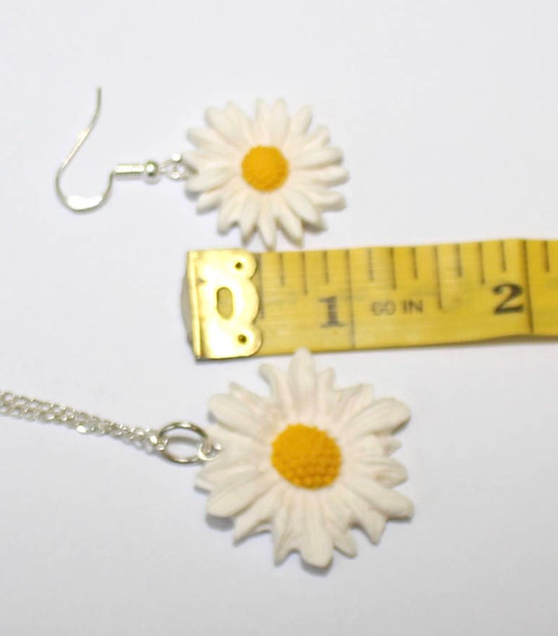May include: A white daisy necklace and earrings set with a yellow centre. The necklace is on a silver chain and the earrings have silver hooks.