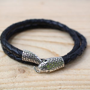 Peut inclure: Bracelet en cuir tressé noir avec un fermoir tête de serpent argenté. Le bracelet est enroulé deux fois et repose sur une surface en bois clair. Le fermoir tête de serpent a un motif d'écailles détaillé.