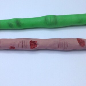 May include: Two novelty pens shaped like fingers. One pen is green and the other is flesh-toned with red painted details.