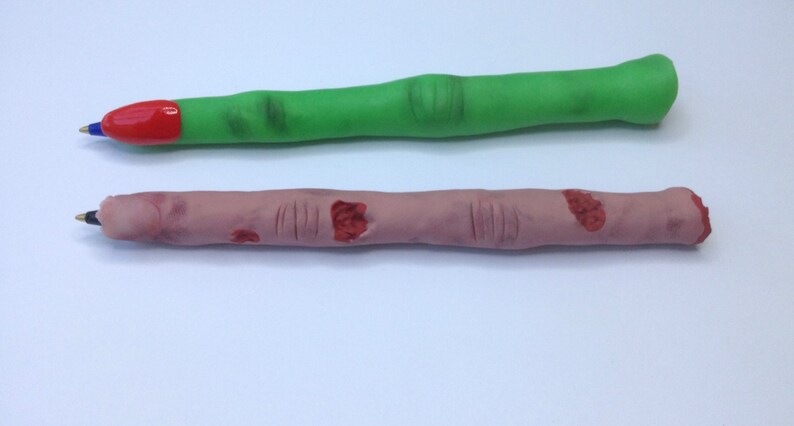 May include: Two novelty pens shaped like fingers. One pen is green and the other is flesh-toned with red painted details.