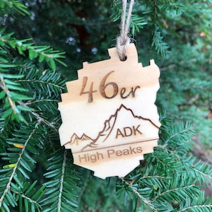 May include: Wooden ornament with the text "46er ADK High Peaks" and a mountain range design. The ornament is light brown and has a natural wood grain. It is hanging from a twine string against a backdrop of green pine needles.