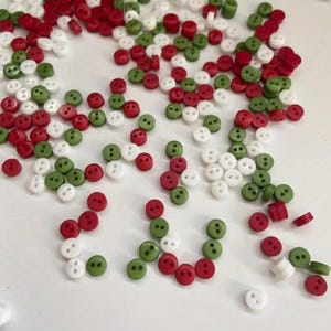 May include: A collection of small, round, two-hole buttons scattered across a surface. The buttons are coloured in red, white, and green, featuring a smooth finish. Each button has two holes in the centre for attachment, with varying orientations.