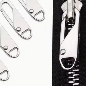 May include: Close-up of silver-coloured zipper pulls and a zipper head. The pulls are elongated with a D-ring at one end. The zipper head is attached to a black fabric.
